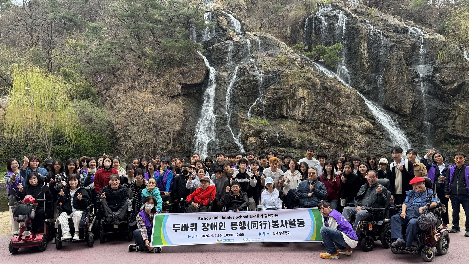 두바퀴 장애인 동행 봉사활동(2026년 4월). 홍제폭포 앞에서 전동휠체어 이용 회원과 봉사 학생들이 현수막을 들고 모였다.
