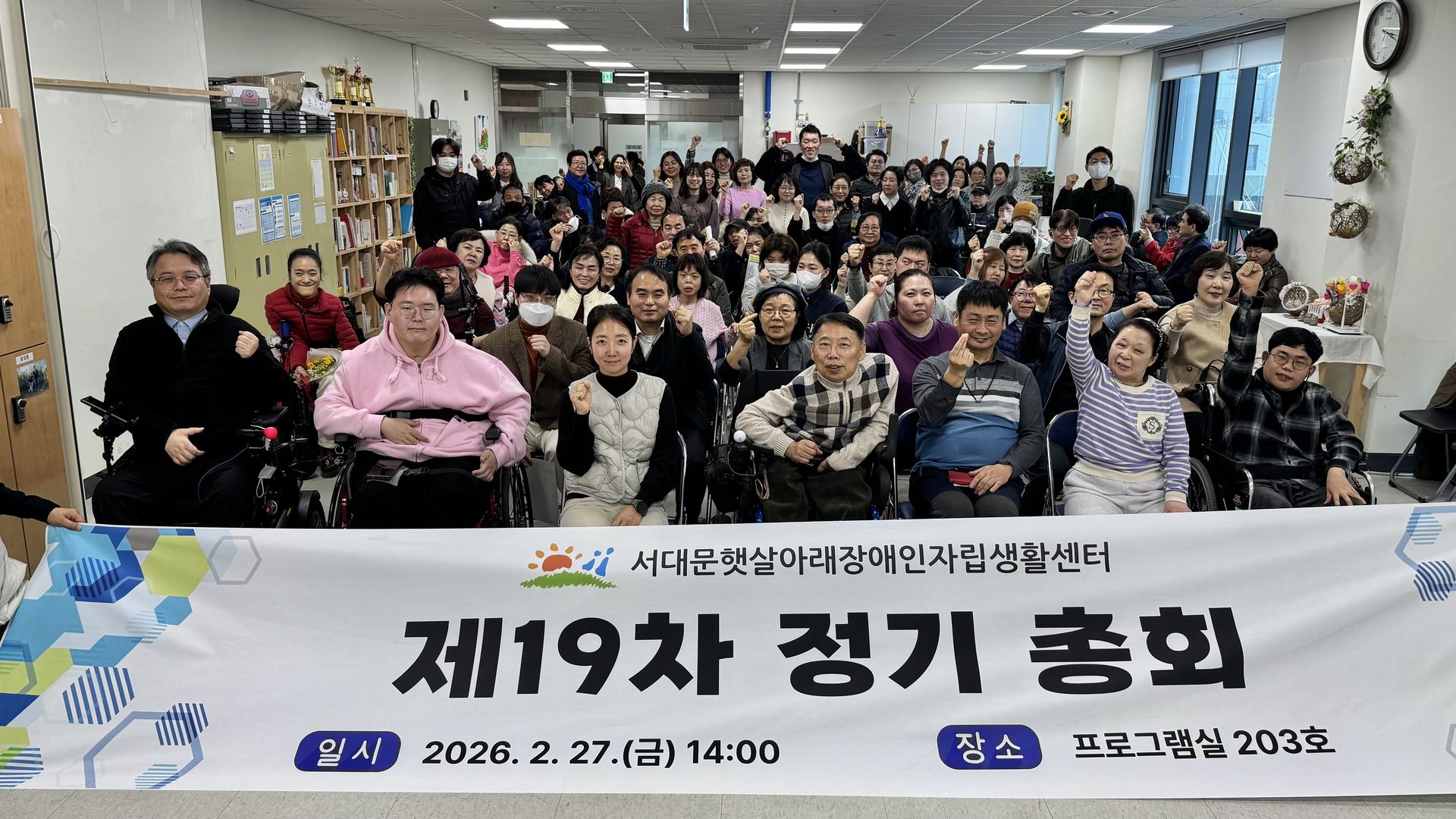 제19차 정기총회(2026년 2월 27일). 회원들이 손을 들어 인사하며 정기총회 현수막 뒤에 모여 있다.