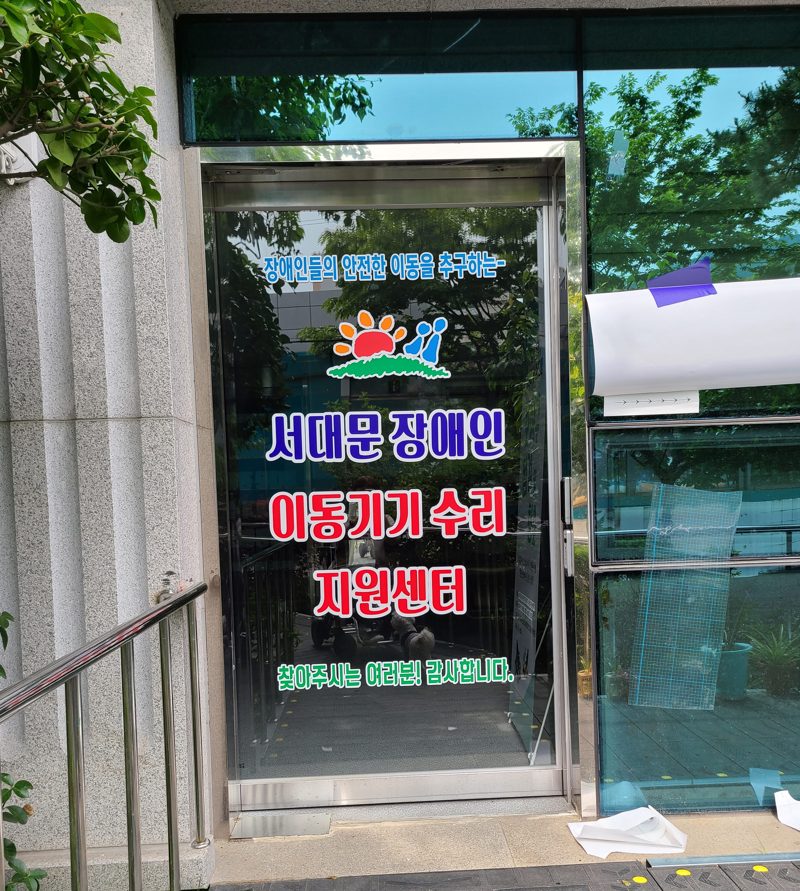 서대문장애인이동기기수리센터 출입문. 유리문에 센터 이름과 로고가 붙어 있고, 입구 옆에 손잡이가 있다.